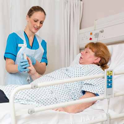  A woman wearing a plastic apron and latex gloves talking to patient lying in a hospital bed